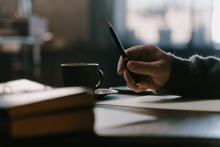 Hand holding a pen above an open book on a desk beside a mug.