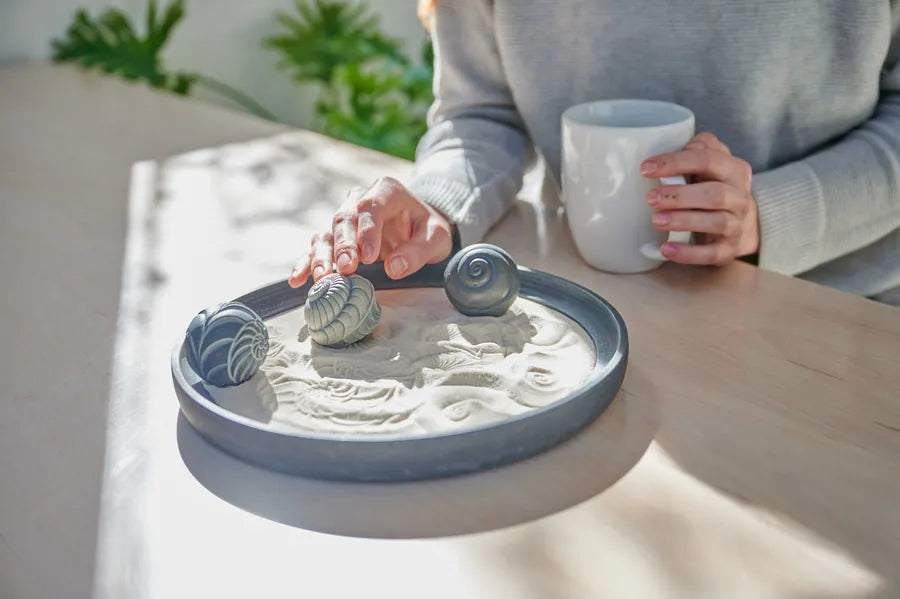 A person holding a coffee mug rolls a spiral-patterned Sand Sphere through a dark tray of sand on a wooden table.