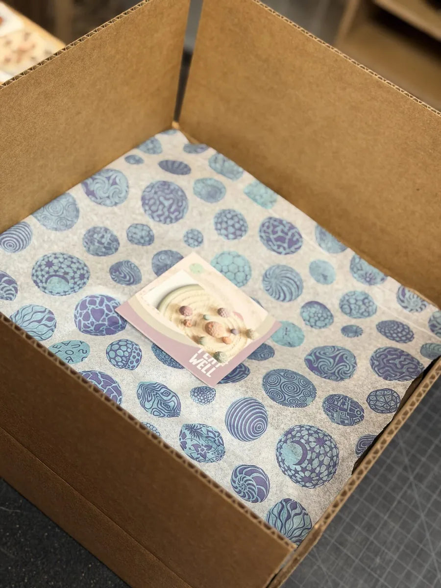 An Olander shipping box lined with printed sphere-pattern tissue, with a Play Well card on top.