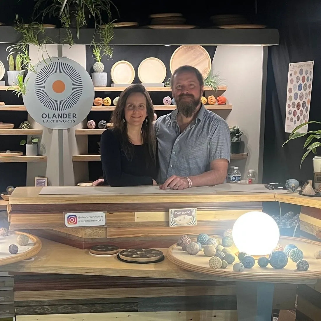Andrew and Sarah Lonnquist behind the Olander Earthworks market booth, with shelves of trays and Sand Spheres, and a lit sand tray to try.