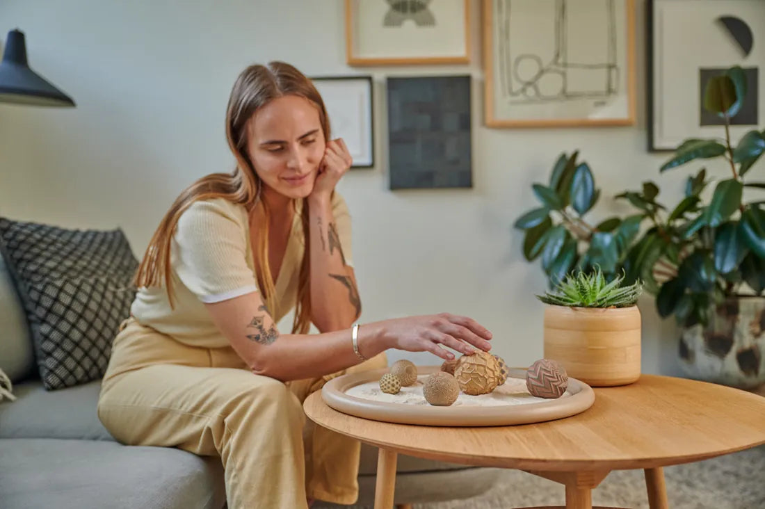 A person leans over a round coffee table to roll a Sand Sphere in a Wonderscape, in a bright living room.