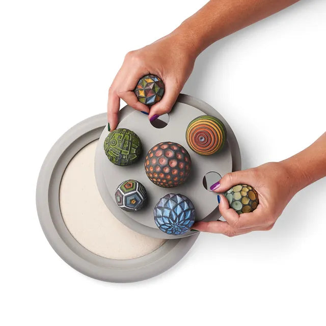 Hands set Sand Spheres into the round openings of a gray tray cover over a Wonderscape tray of sand.