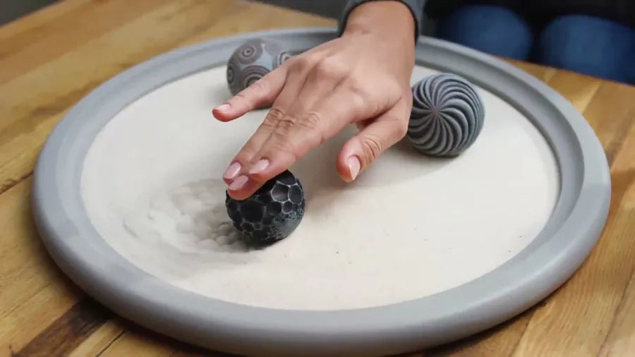 A hand starts a fresh track with a textured sphere in smoothed sand in a gray tray.