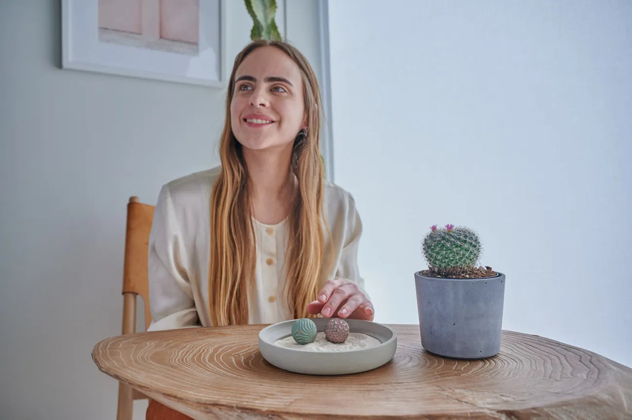 A smiling person sits at a round wooden table beside a gray Wonderscape tray with two patterned Sand Spheres and a potted cactus.