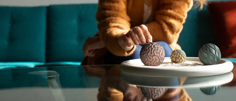 A hand rolls a patterned Sand Sphere in a white Wonderscape on a glass coffee table.