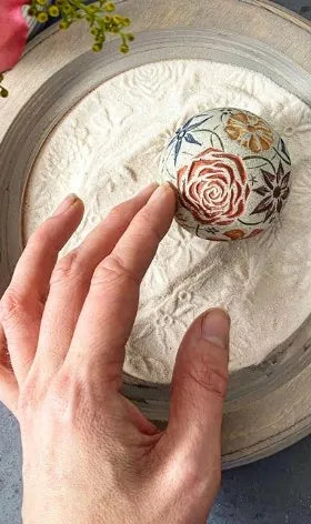A hand rolls a Sand Sphere carved with flowers through pale sand, leaving flower prints.