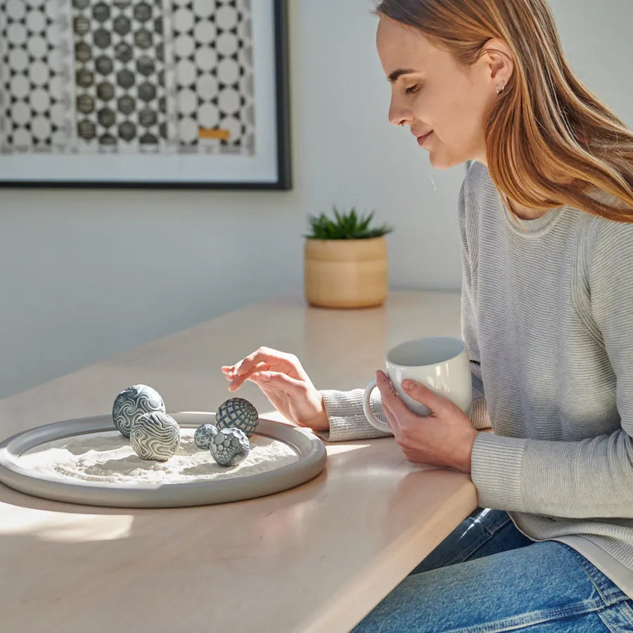 A person at a desk holds a mug and rolls Sand Spheres in a Wayfinder Wonderscape, a wide gray tray.